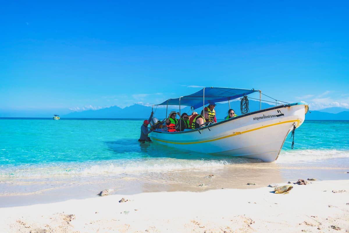 Tourists wearing life jackets board a small motorboat on a tropical beach with clear turquoise water and distant mountains.