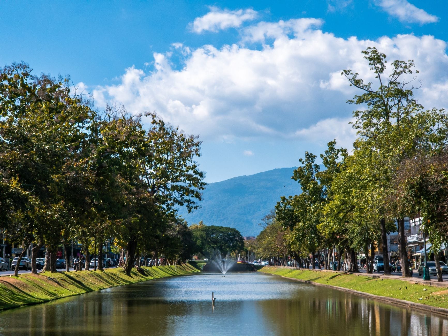 Cảnh hào thành của thành phố Chiang Mai được viền bởi những hàng cây, đài phun nước ở trung tâm và những ngọn núi xa xa dưới bầu trời xanh.