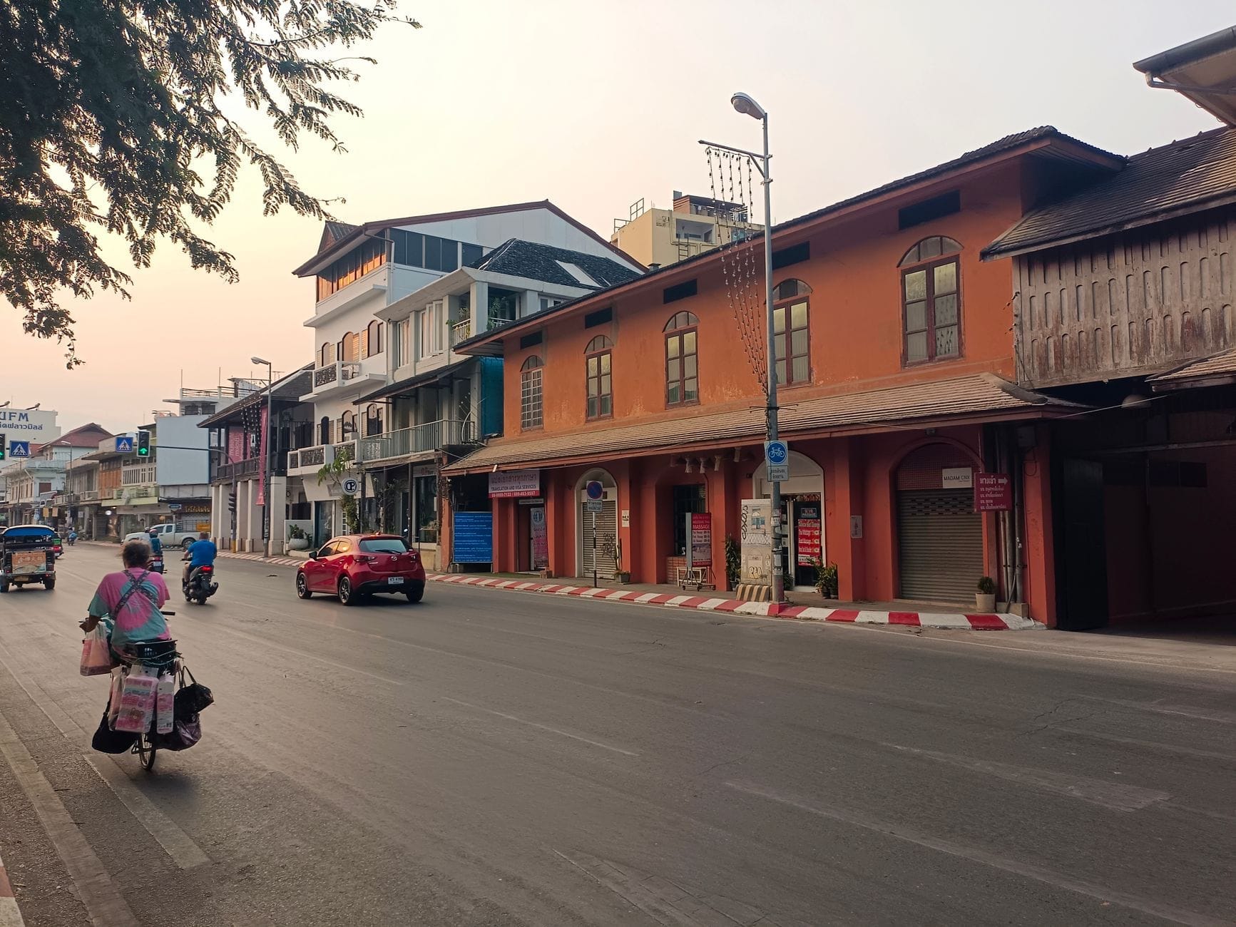 Escena callejera de Chiang Mai con motos, un auto rojo y personas en un tuk-tuk pasando frente a coloridos edificios en una tranquila mañana.