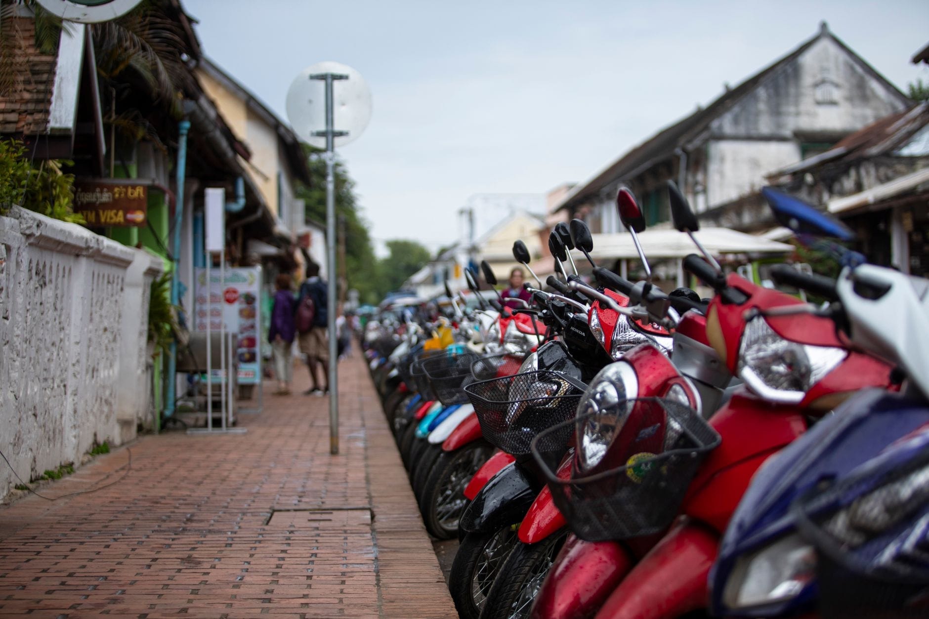 Hàng xe scooter đậu dọc theo vỉa hè trong một thành phố Đông Nam Á với các tòa nhà và biển hiệu đường phố hiện ra rõ ràng.