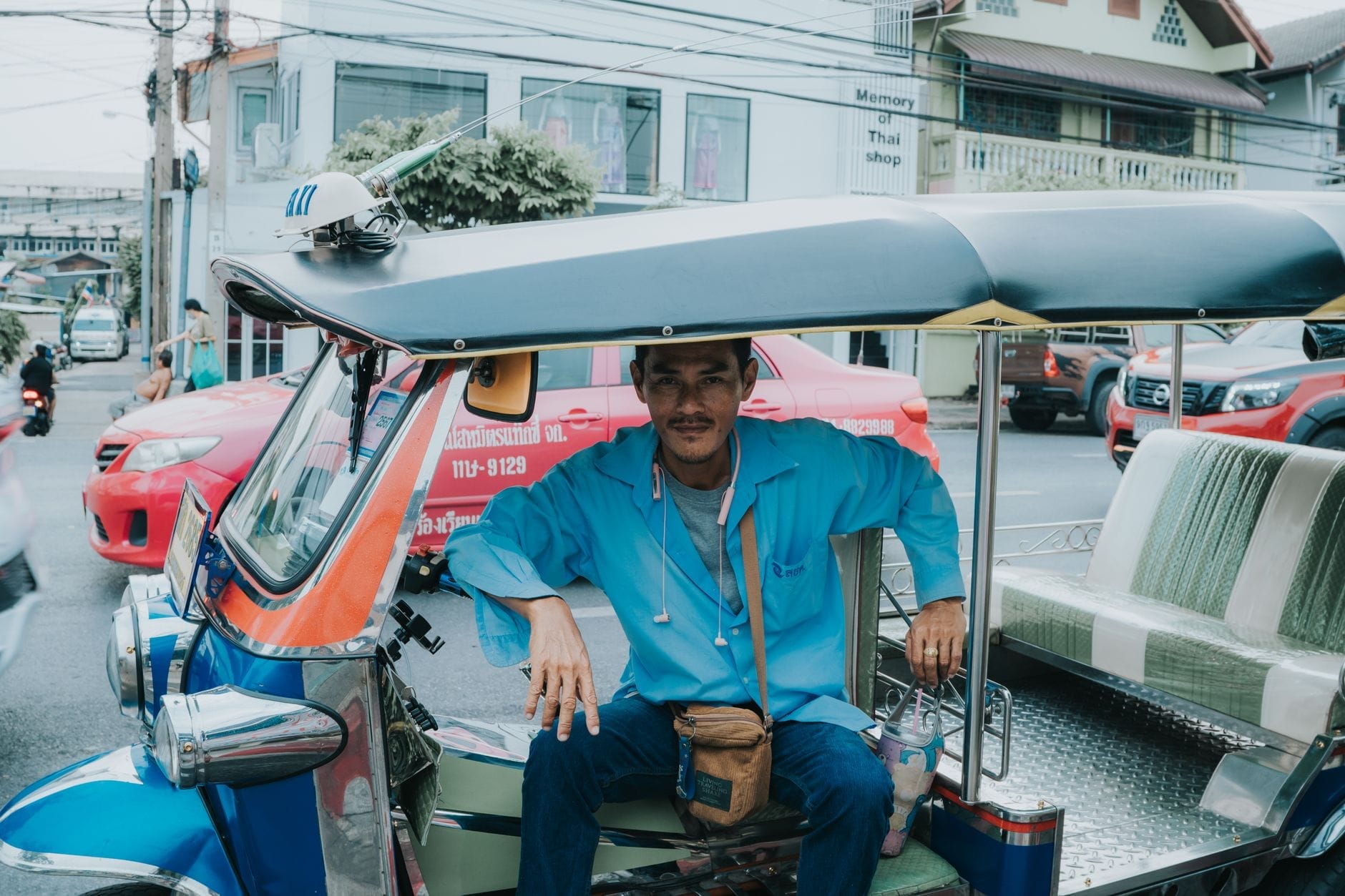 Một tài xế tuk-tuk ngồi trong chiếc tuk-tuk màu xanh trên đường phố Chiang Mai với các tòa nhà đô thị và giao thông ở phía sau.