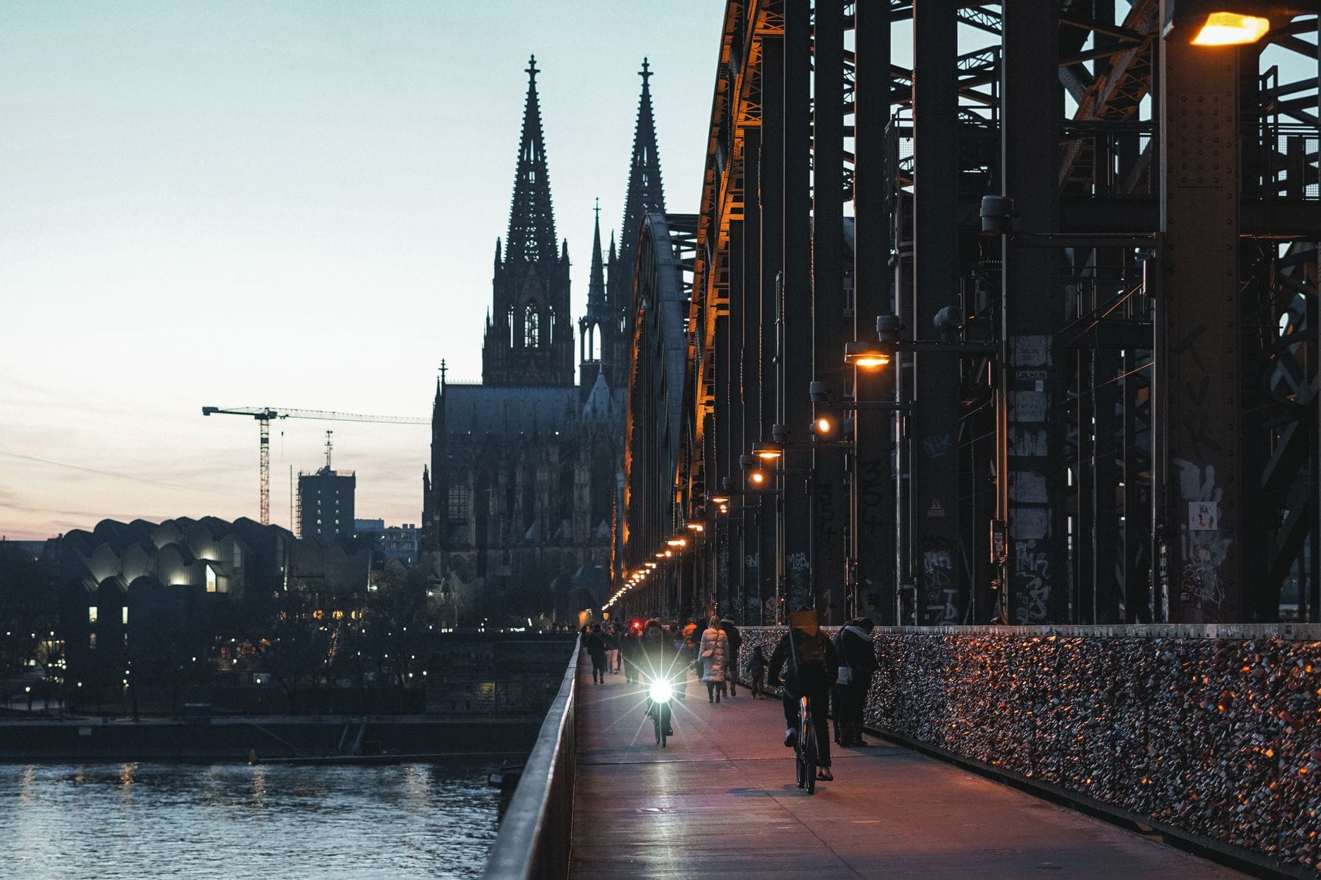ผู้คนเดินและขี่จักรยานบนสะพาน Hohenzollern โดยมีมหาวิหาร Cologne และเส้นขอบฟ้าของเมืองเป็นฉากหลังในยามพลบค่ำ