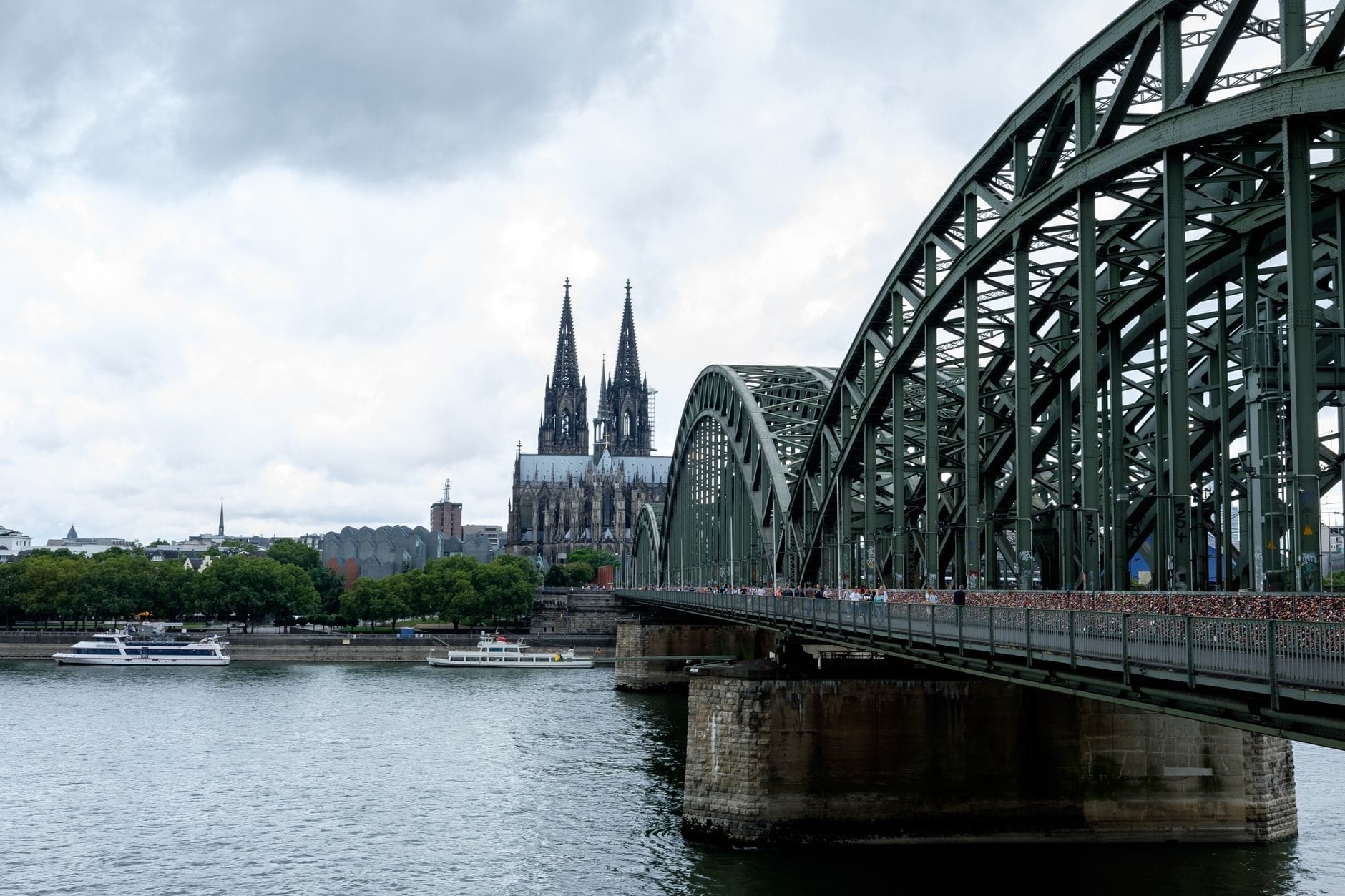 ภาพกว้างของสะพาน Hohenzollern เมืองโคโลญจน์ข้ามแม่น้ำไรน์ มีรถไฟ คนเดิน และโบสถ์โคโลญจน์อยู่พื้นหลังในวันที่เมฆครึ้ม