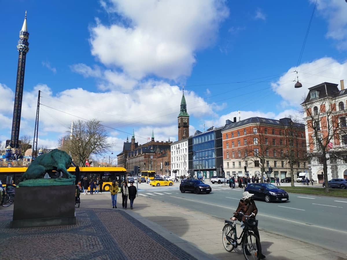 Belebte Straße in Kopenhagen mit Fußgängern, Radfahrern, Autos, Bussen und Stadtgebäuden unter einem teilweise bewölkten Himmel.
