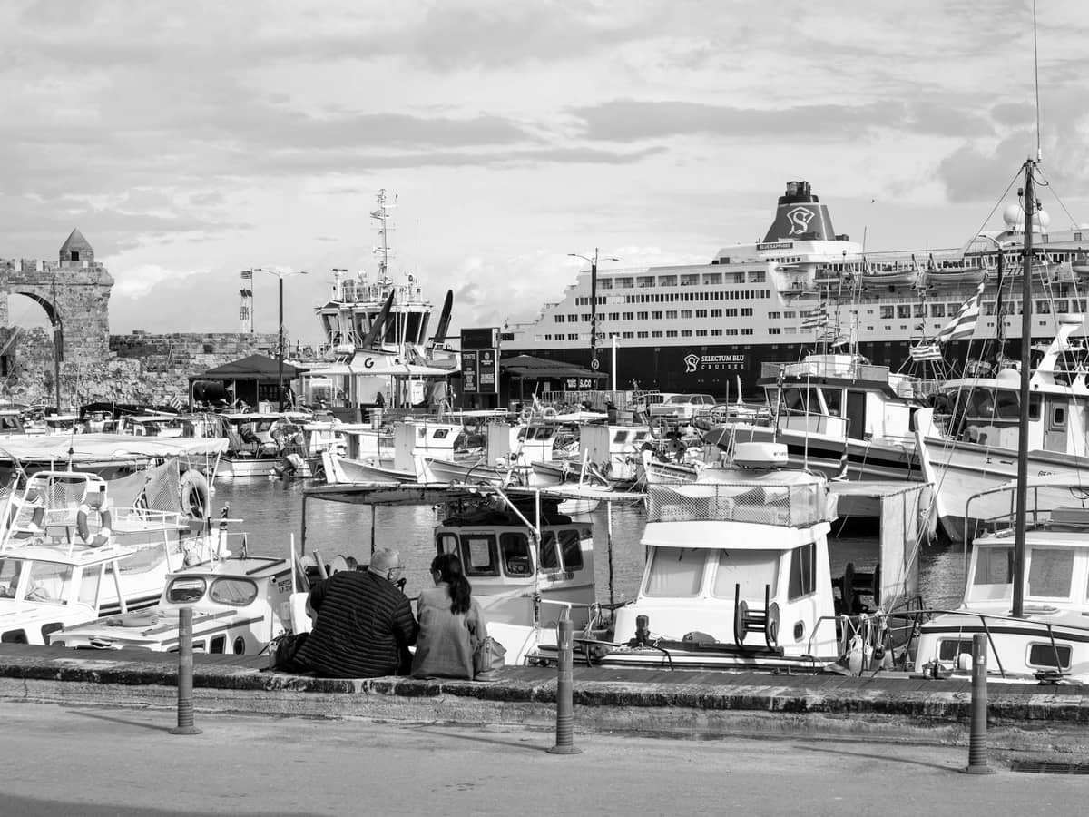 Porto di Heraklion a Creta con traghetti ormeggiati, barche da pesca e persone sedute lungo il lungomare, che illustrano le opzioni di trasporto in traghetto.