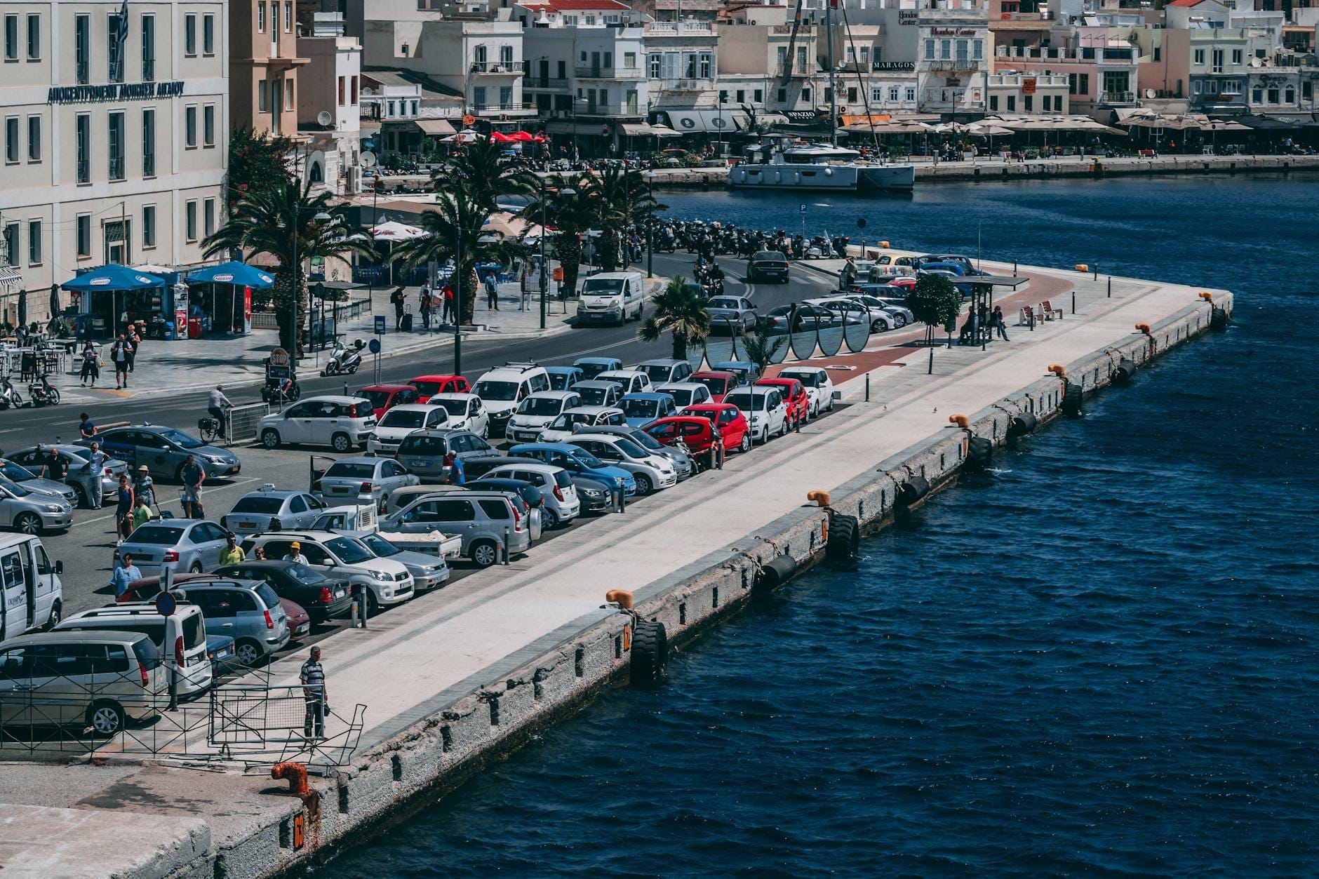 Um grande estacionamento cheio de carros ao lado de um pitoresco porto de Creta, com prédios da cidade e palmeiras ao longo do calçadão à beira-mar.