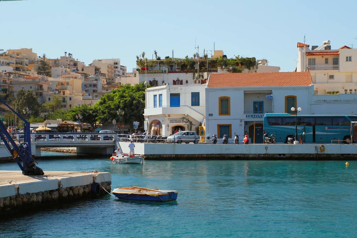 Scena sul lungomare di Creta con un autobus interurbano KTEL blu parcheggiato vicino a un porto, circondato da edifici mediterranei.