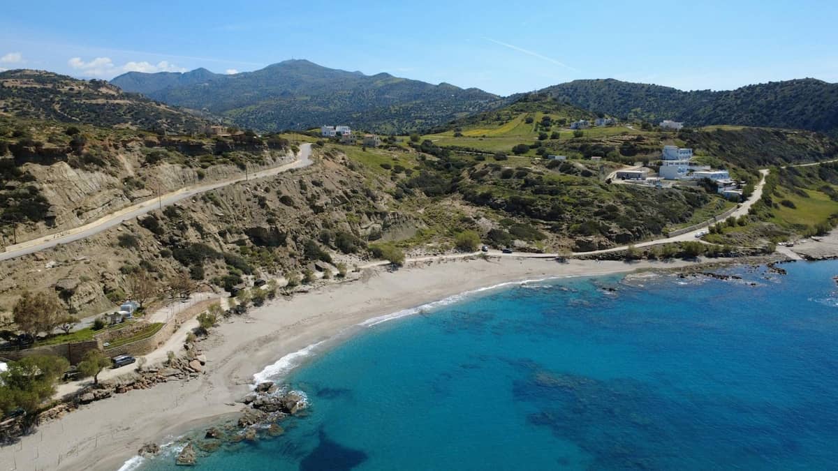 Una tortuosa strada costiera lungo colline aspre e una spiaggia, con edifici sparsi e montagne in lontananza a Creta.