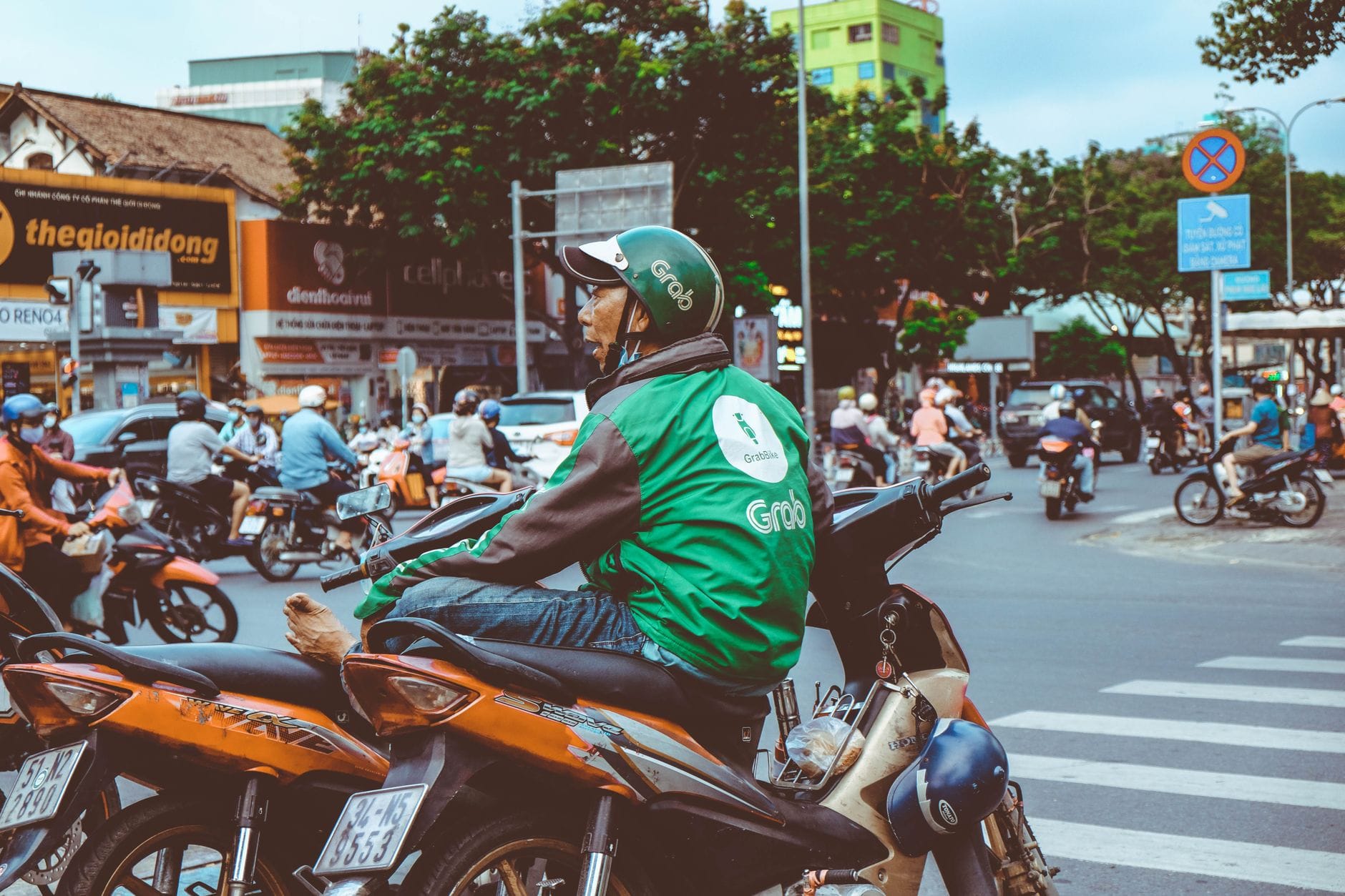 คนขับมอเตอร์ไซค์แท็กซี่ Grab สวมชุดสีเขียวรอที่สี่แยกในเมืองดานังที่ยุ่งวุ่นวาย ล้อมรอบด้วยรถมอเตอร์ไซค์คันอื่นและความเคลื่อนไหวบนท้องถนน