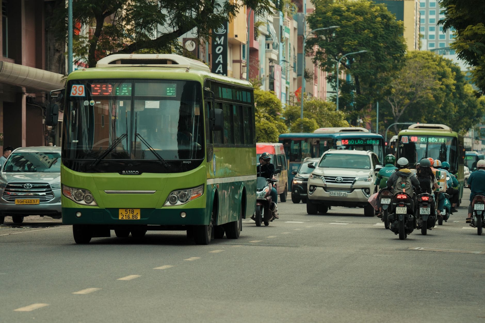 รถเมล์สีเขียวและมอเตอร์ไซค์บนถนนในเมืองที่พลุกพล่านในเวียดนาม พร้อมอาคารและต้นไม้เรียงรายสองข้างทาง