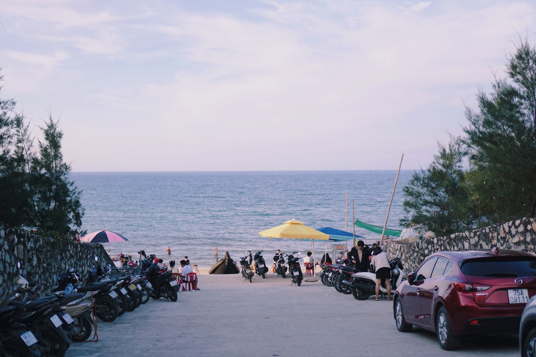 แถวของมอเตอร์ไซค์จอดและรถยนต์บางคันที่นำไปตามทางคอนกรีตสู่ชายหาดทรายที่คึกคักพร้อมร่มและผู้คนใกล้ทะเล