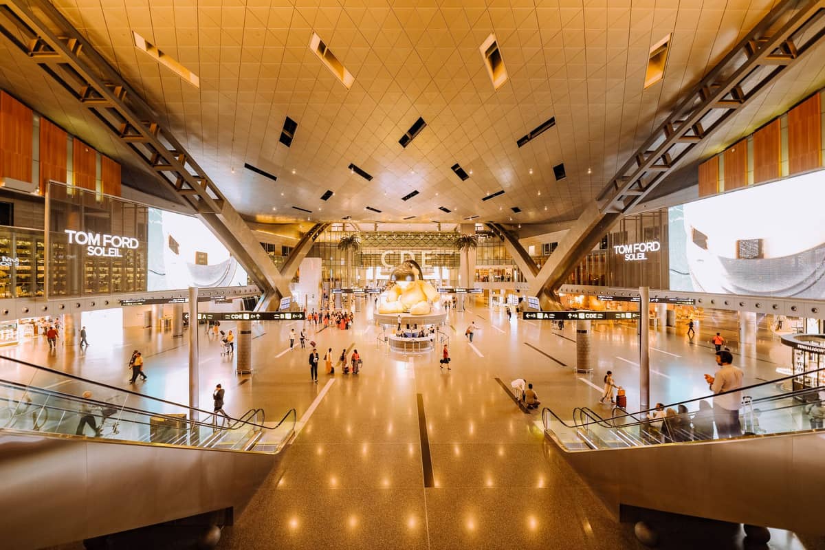 Weitläufige Ansicht der hell erleuchteten Haupthalle des Hamad International Airport mit Reisenden, Rolltreppen und der ikonischen gelben Teddybär-Skulptur.
