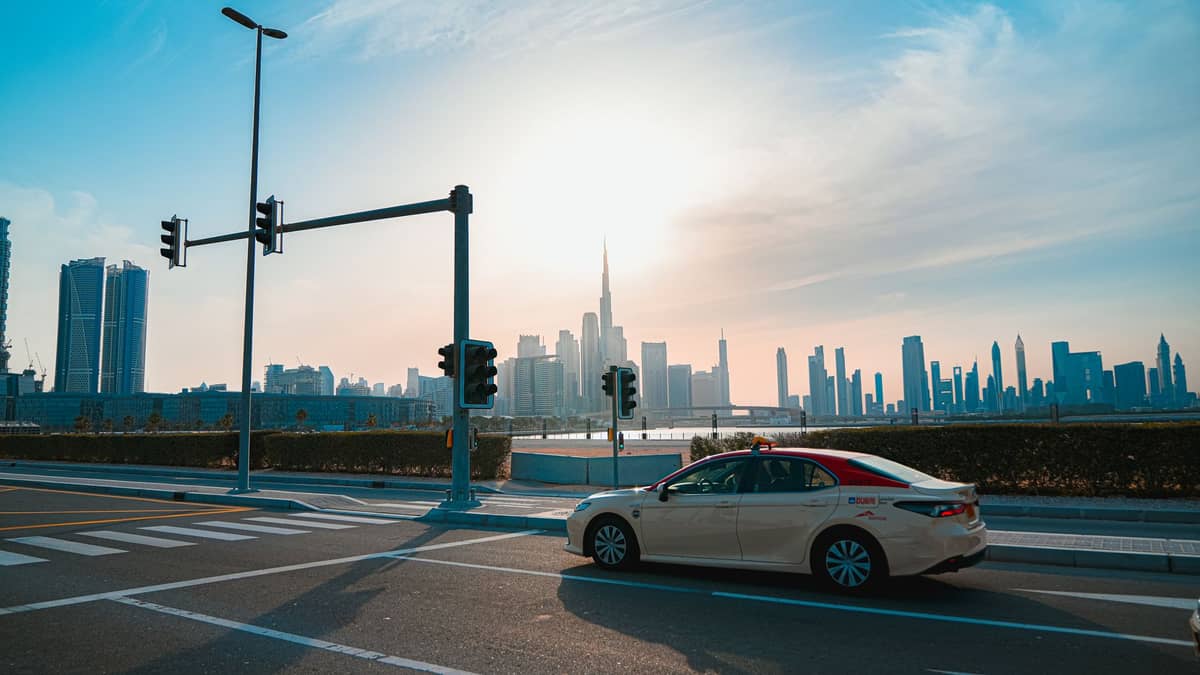 Cremefarbenes Dubai-RTA-Taxi fährt mit dem Burj Khalifa und der Skyline von Dubai im Hintergrund bei Sonnenuntergang.