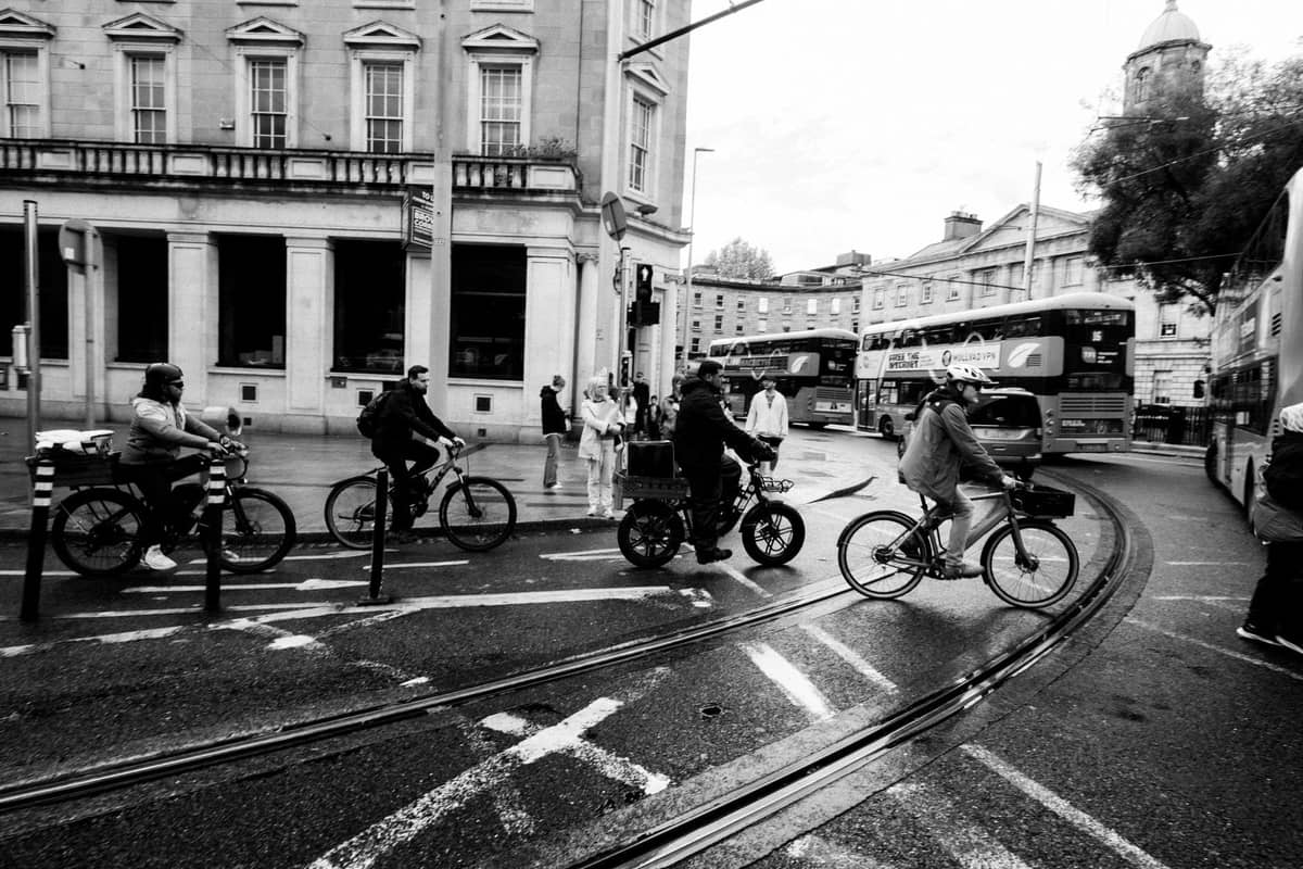 Radfahrer an einer belebten Kreuzung in Dublin mit sichtbaren Straßenbahnschienen, Bussen und historischen Gebäuden im Hintergrund.