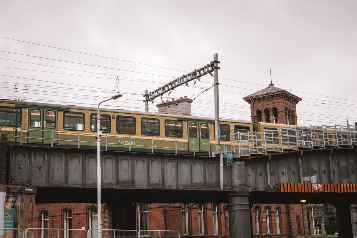 Ein DART-Zug überquert eine erhöhte Eisenbahnbrücke in Dublin, mit historischen Backsteingebäuden und einem Uhrturm im Hintergrund.