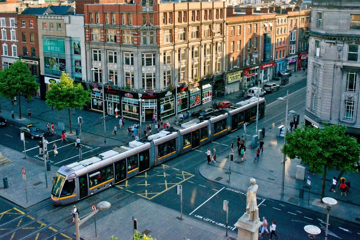 Eine Luas-Straßenbahn fährt durch eine lebhafte Straße in Dublin mit historischen Gebäuden, geschäftigen Fußgängern und städtischem Verkehr an einer wichtigen Kreuzung.