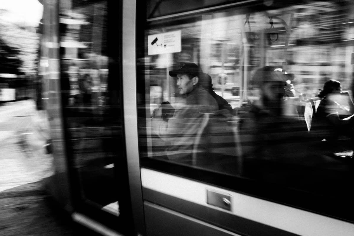 Ein Mann sitzt in einer modernen Straßenbahn, deren Fenster die Stadtstraße draußen spiegeln, aufgenommen in Schwarz-Weiß in Dublin.