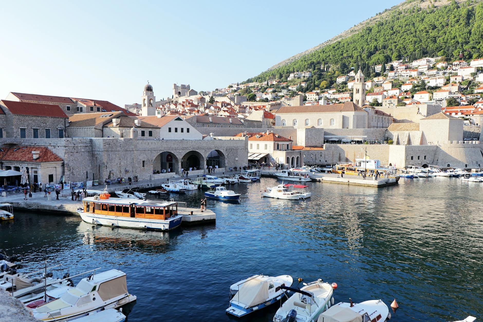 Bateaux et ferries amarrés au Vieux-Port de Dubrovnik avec les remparts de la ville et des bâtiments historiques en arrière-plan sous la lumière du soleil.