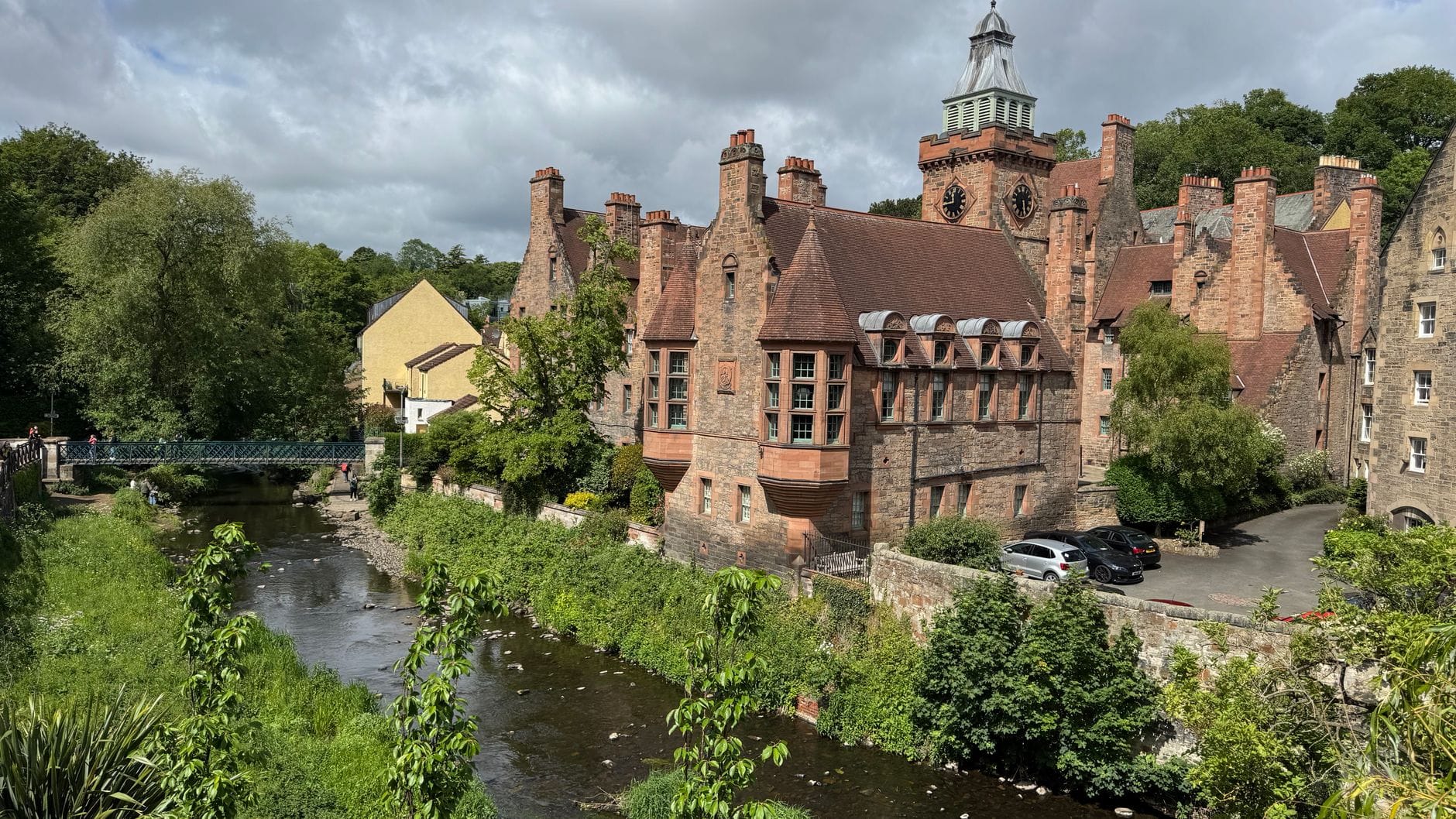Landschaftsansicht von Dean Village in Edinburgh mit dem Water of Leith, historischen Steingebäuden, üppigem Grün und einer Fußgängerbrücke im Sommerlicht.