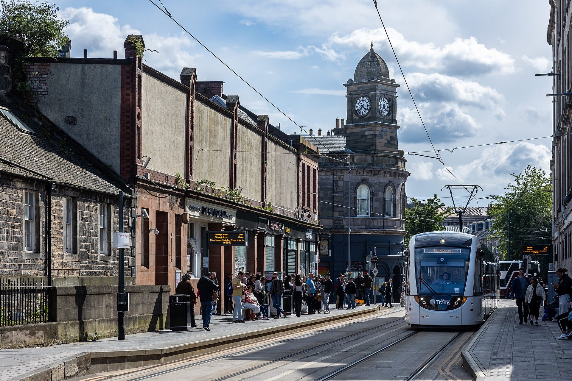 Moderne Edinburgh-Straßenbahn hält an einer belebten Straßenstation mit wartenden Menschen, Oberleitungen und einem historischen Uhrenturm im Hintergrund.