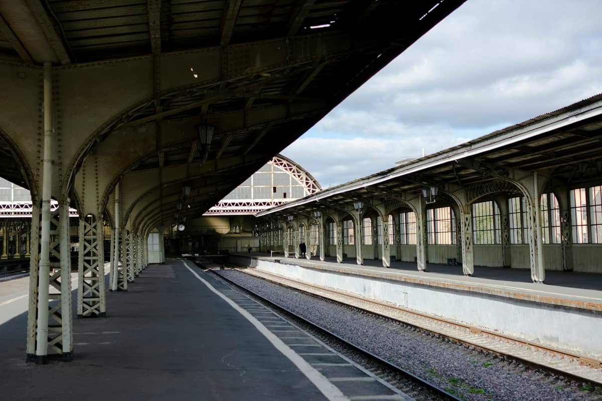 View of covered train platforms and ironwork at a classic European railway station under a cloudy sky.