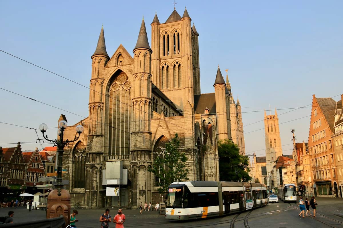 Modern tram passes in front of St. Nicholas' Church in central Ghent with historic architecture in the evening sunlight.