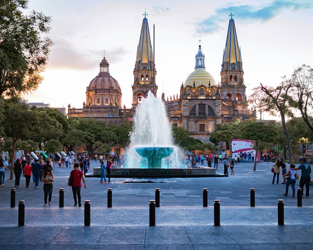 Menschen, die bei Sonnenuntergang über einen belebten Platz mit einem Brunnen und der Kathedrale von Guadalajara im Hintergrund spazieren.
