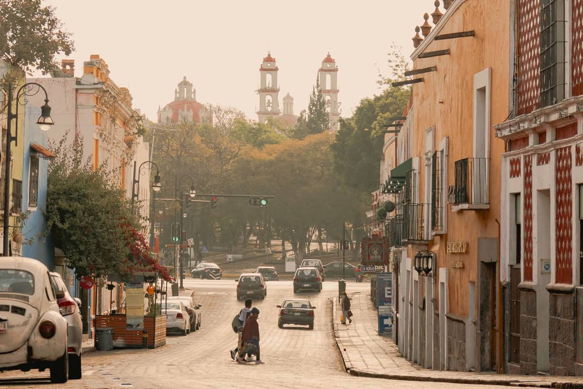 Straßenszene in Guadalajara mit mehreren Autos, darunter klassische Volkswagen Beetles, bunten Gebäuden und Kirchtürmen im Hintergrund.
