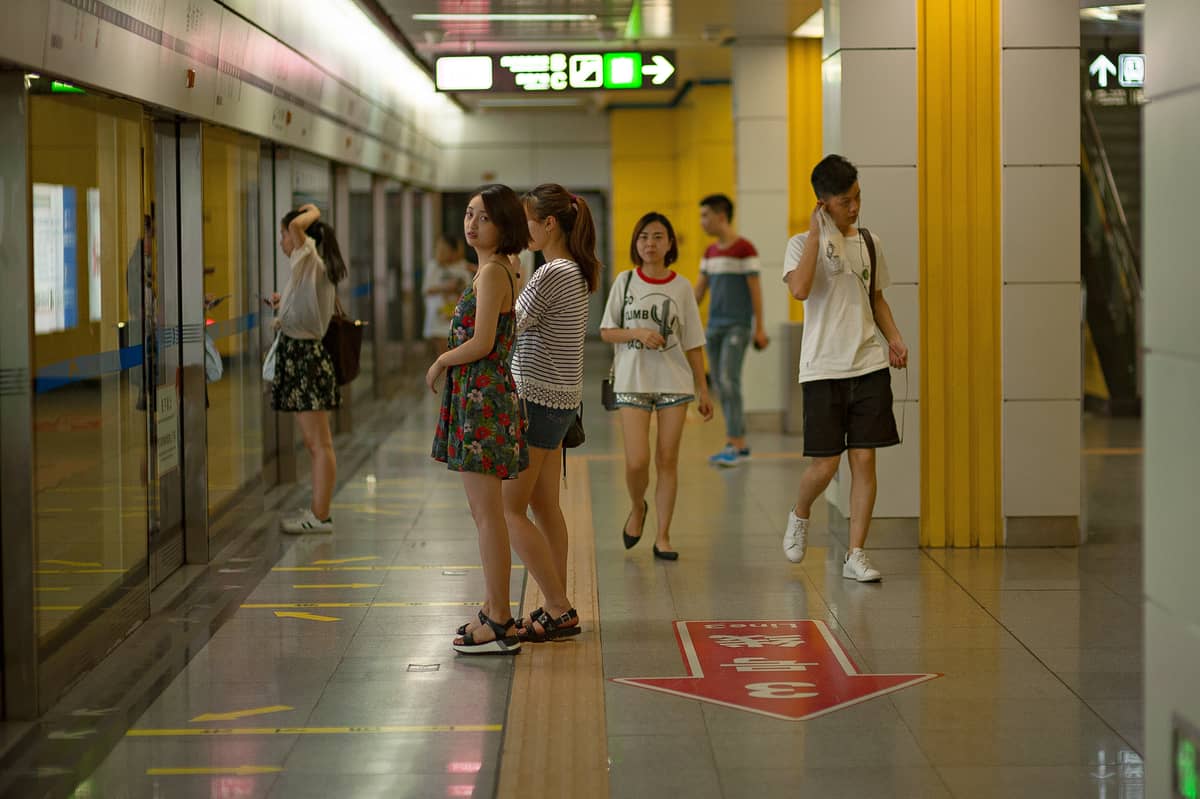 Passengers wait on a clean, modern platform of the Guangzhou Metro, with yellow pillars, glass safety doors, and bilingual signage overhead.