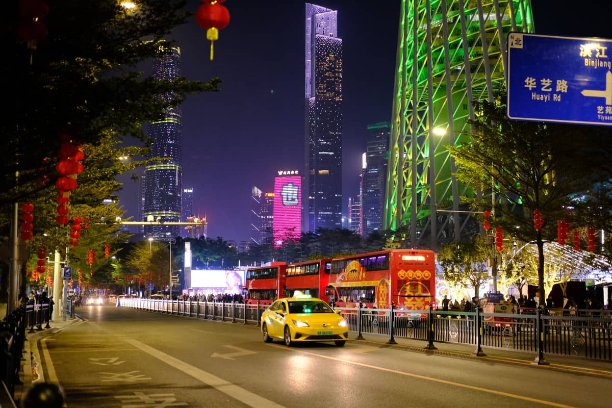 Yellow taxi driving along a brightly lit Guangzhou street at night near modern skyscrapers and Canton Tower.