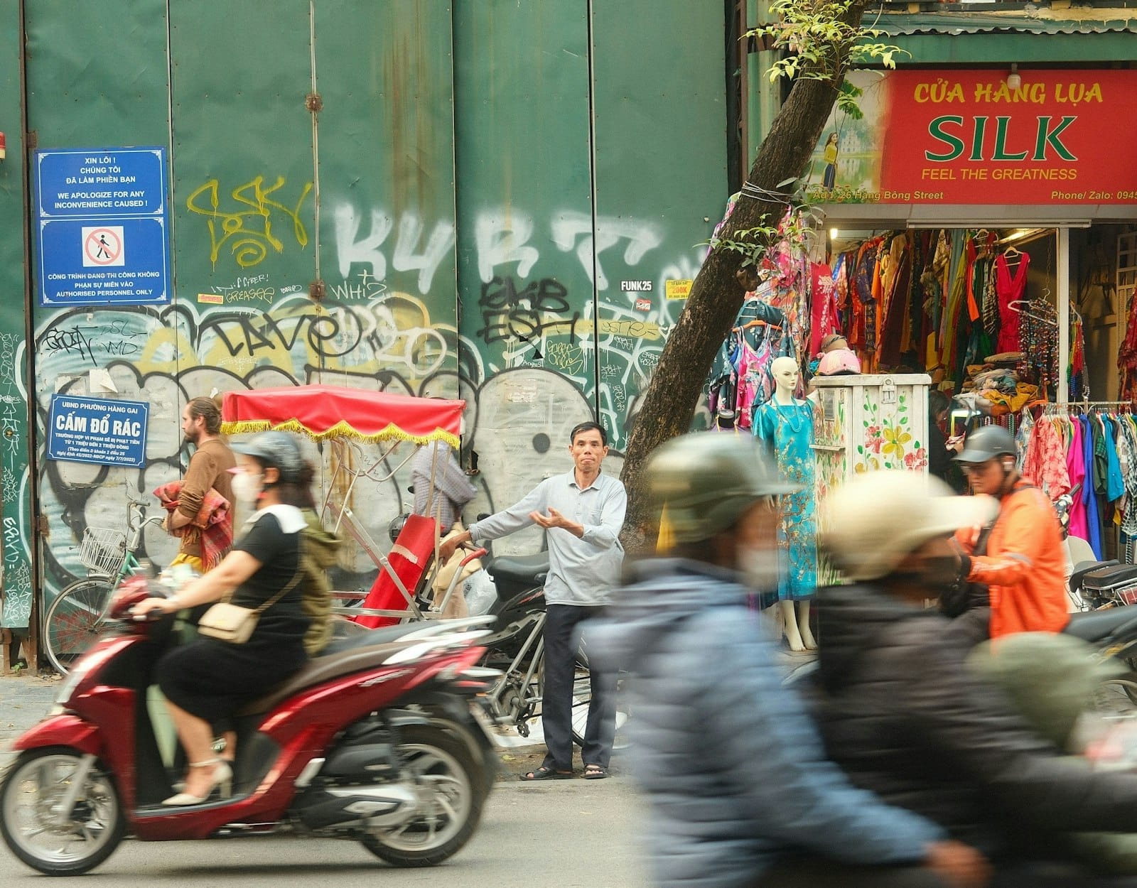 Busy Hanoi street scene with people riding motorbikes and a cyclo in front of local shops and colorful storefronts.