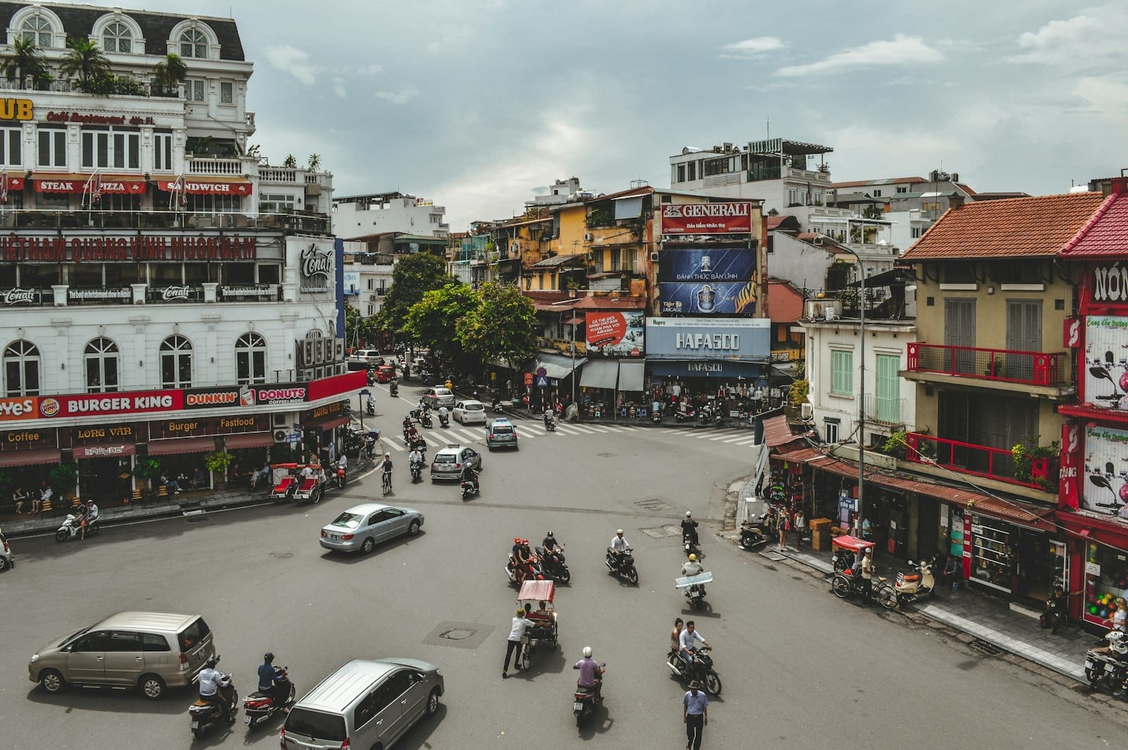 Busy street intersection in Hanoi with cars, motorbikes, cyclos, and pedestrians navigating between colorful buildings and shops on a cloudy day.