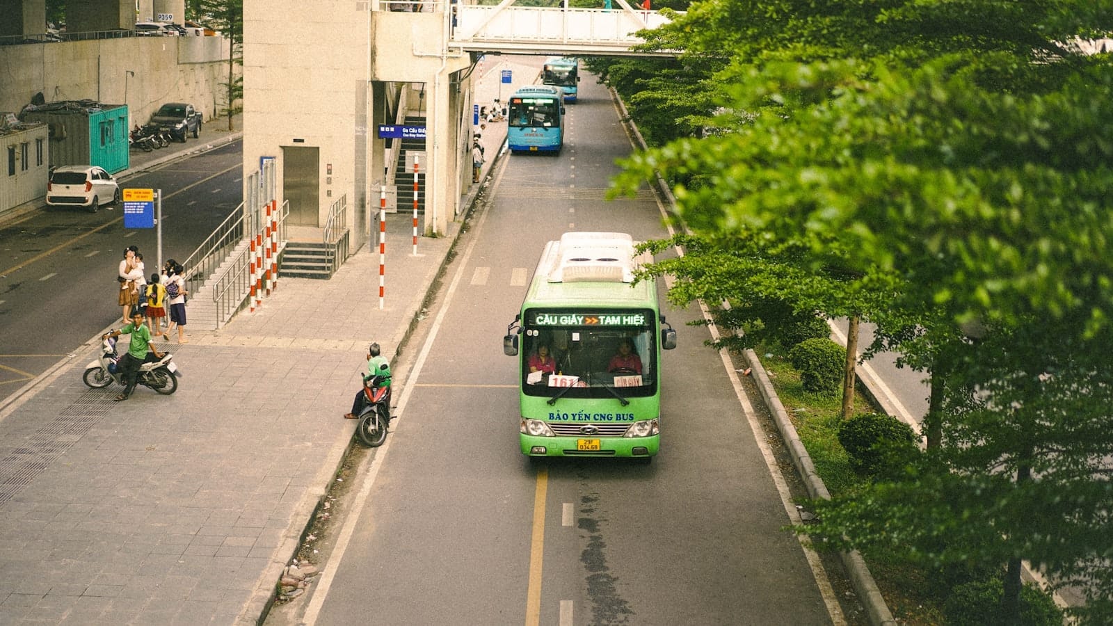 Green public bus driving down a street in Hanoi with another bus in the distance, surrounded by trees and buildings.