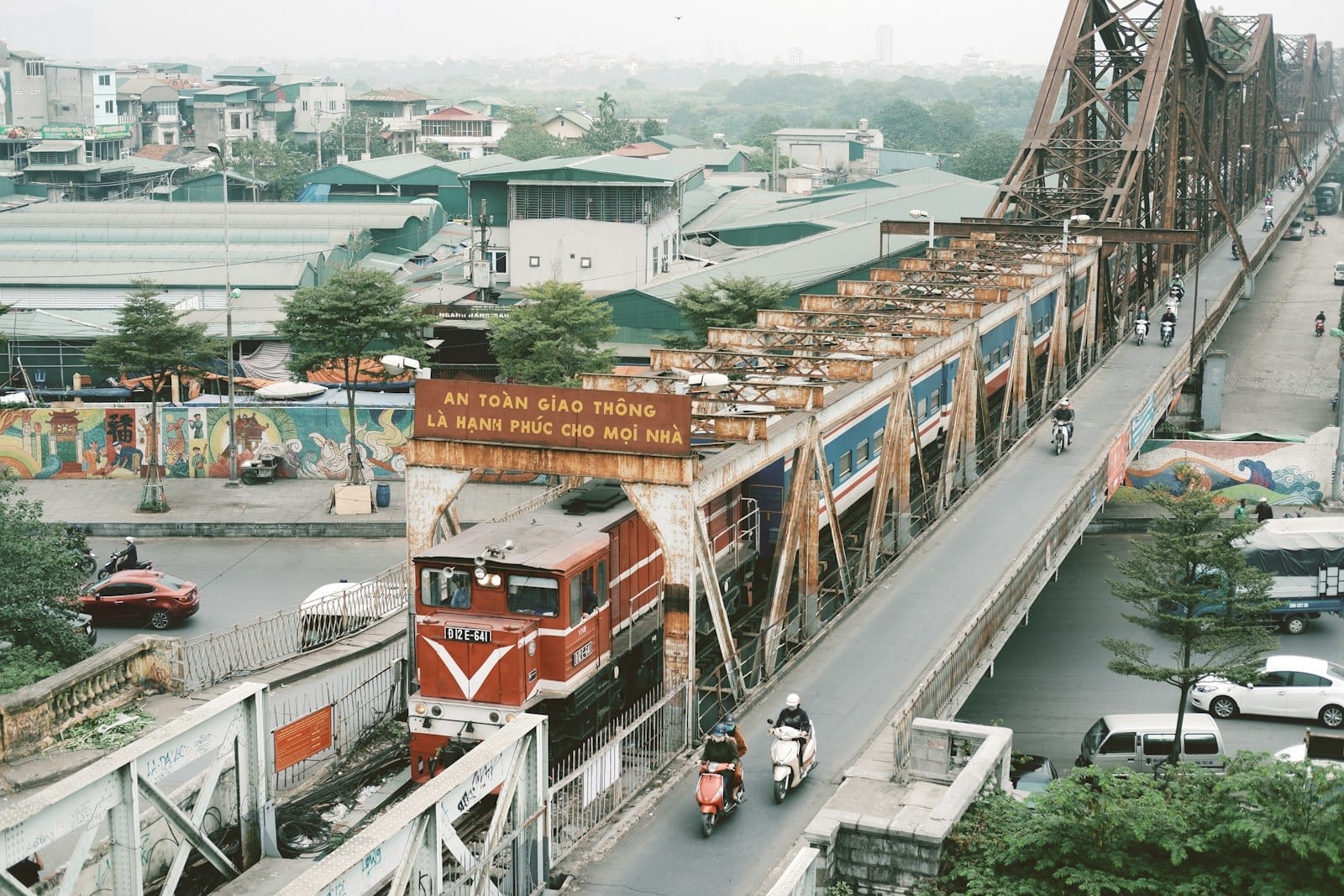 A passenger train crossing Hanoi's iconic Long Bien Bridge with city buildings and motorbikes below, illustrating departure routes for day trips.