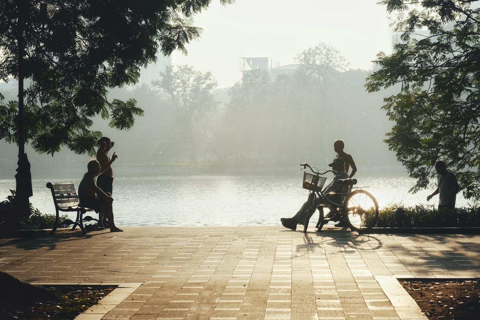 People walking, sitting, and cycling along a paved lakeside path shaded by trees, with misty light over the water in Hanoi.
