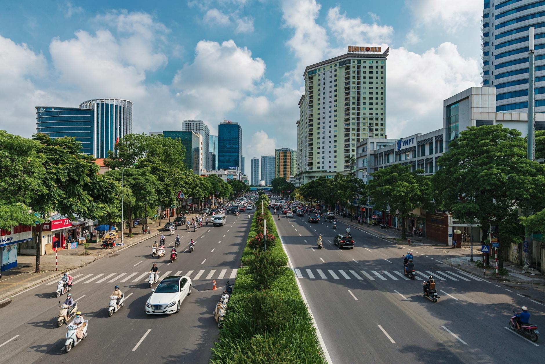 Wide boulevard in Hanoi with cars and motorbikes moving between districts, surrounded by modern buildings and trees under a bright sky.