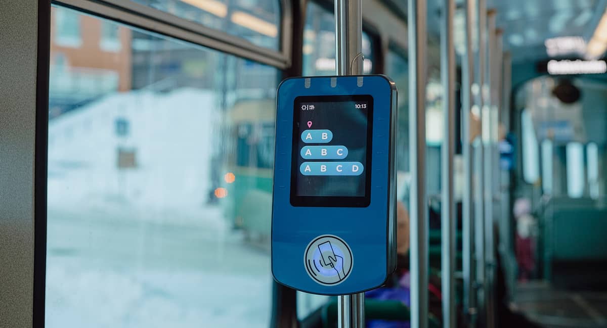 Um leitor de cartão HSL azul num elétrico de Helsínquia a exibir as opções de zonas ABCD, com o interior do elétrico e uma rua nevada visível lá fora.