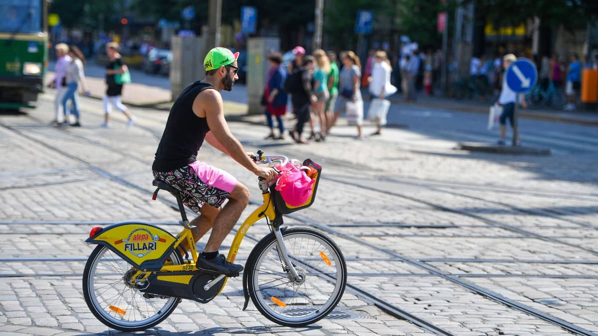 Homem andando de bicicleta amarela de Helsinki em rua de paralelepípedos com trilhos de bonde, pedestres e cena urbana ao fundo.