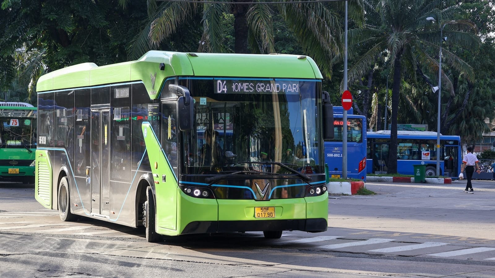 Một chiếc xe buýt công cộng màu xanh lá tại Thành phố Hồ Chí Minh ở một ngã tư đường với các xe buýt khác và cây cọ ở phía sau.