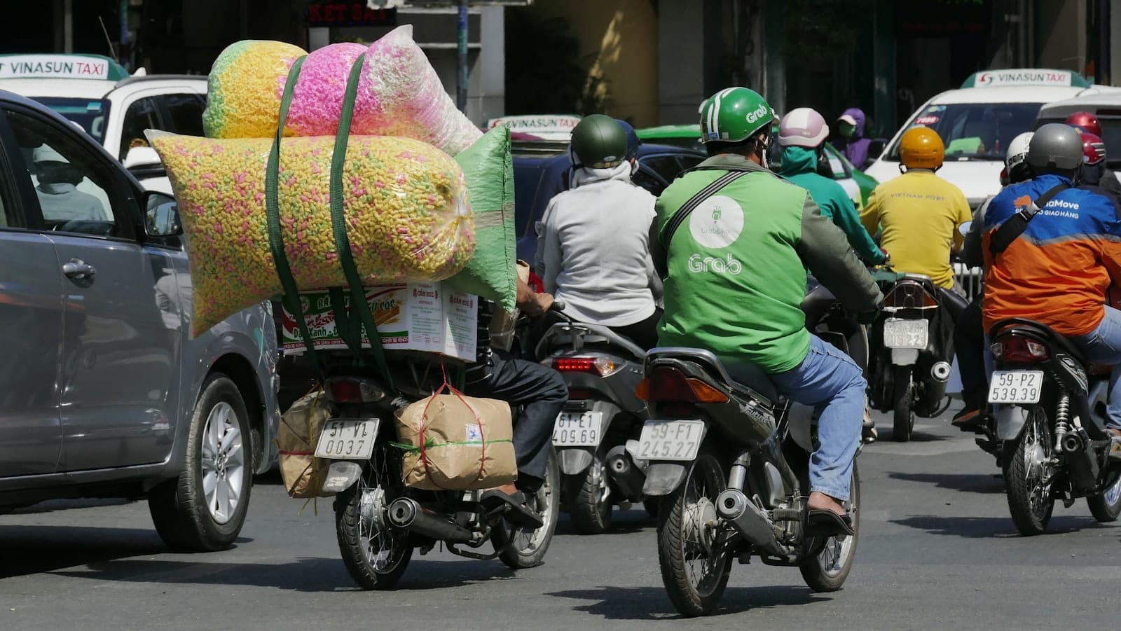 Nhiều người lái xe máy, trong đó có tài xế Grab mặc áo khoác xanh lá và đội mũ bảo hiểm, cùng các taxi trong dòng xe trên một con phố đông đúc ở Thành phố Hồ Chí Minh.