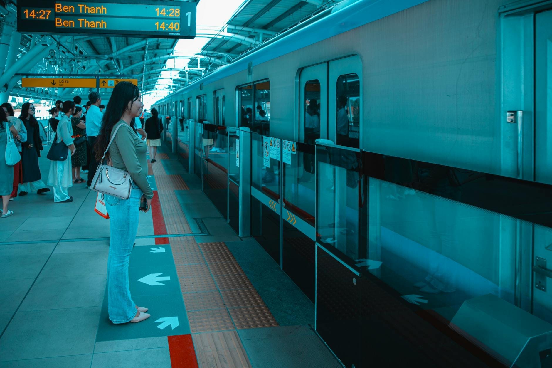 Hành khách đứng trên sân ga tàu điện ngầm hiện đại ở Thành phố Hồ Chí Minh, chờ tàu đến ga Bến Thành, với biển chỉ dẫn rõ ràng phía trên.