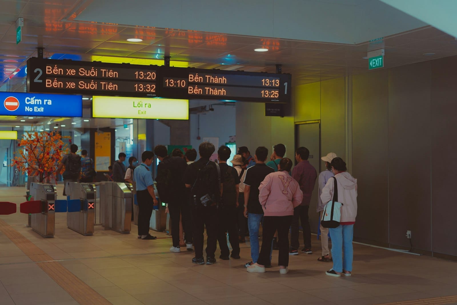 Hành khách xếp hàng tại một ga tàu điện ngầm hoặc đường sắt được chiếu sáng rực rỡ ở Thành phố Hồ Chí Minh, với các biển chỉ dẫn đến ga Suối Tiên và Bến Thành.