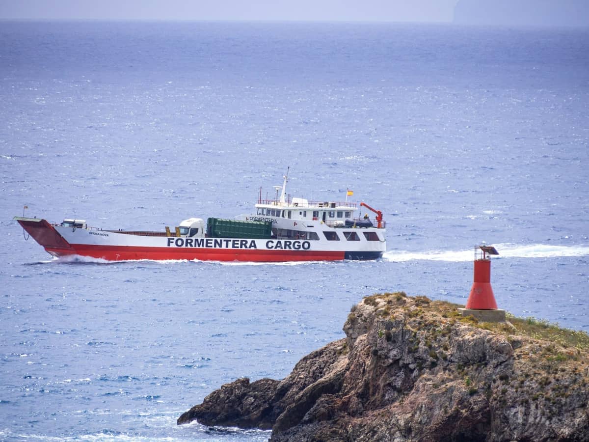 A ferry labeled 'Formentera Cargo' sails past a rocky headland with a red navigation beacon on the coast near Ibiza.