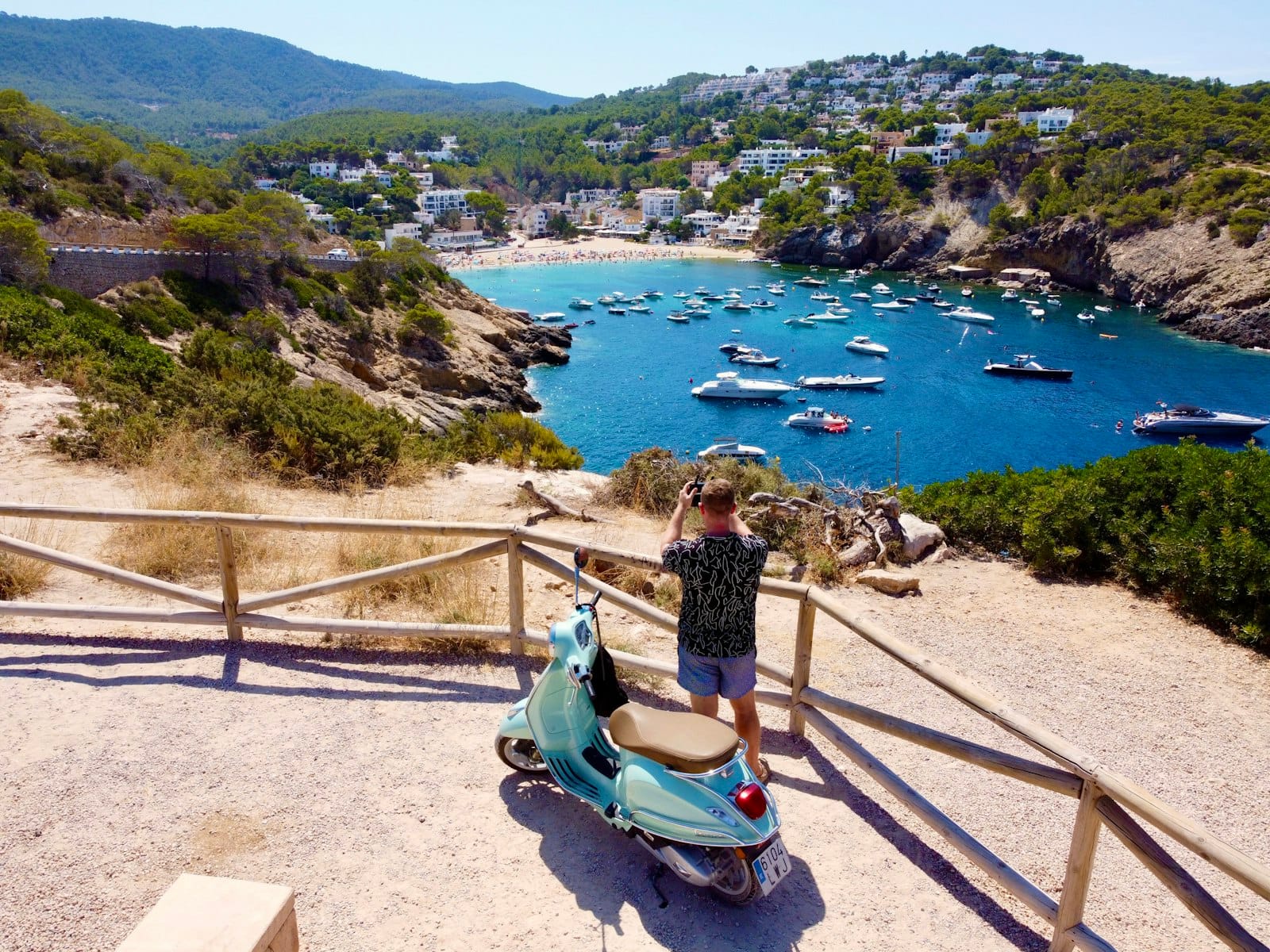 Pessoa com uma scooter contempla uma baía panorâmica de Ibiza com barcos, pinheiros, colinas e uma vila ao fundo sob um céu limpo.