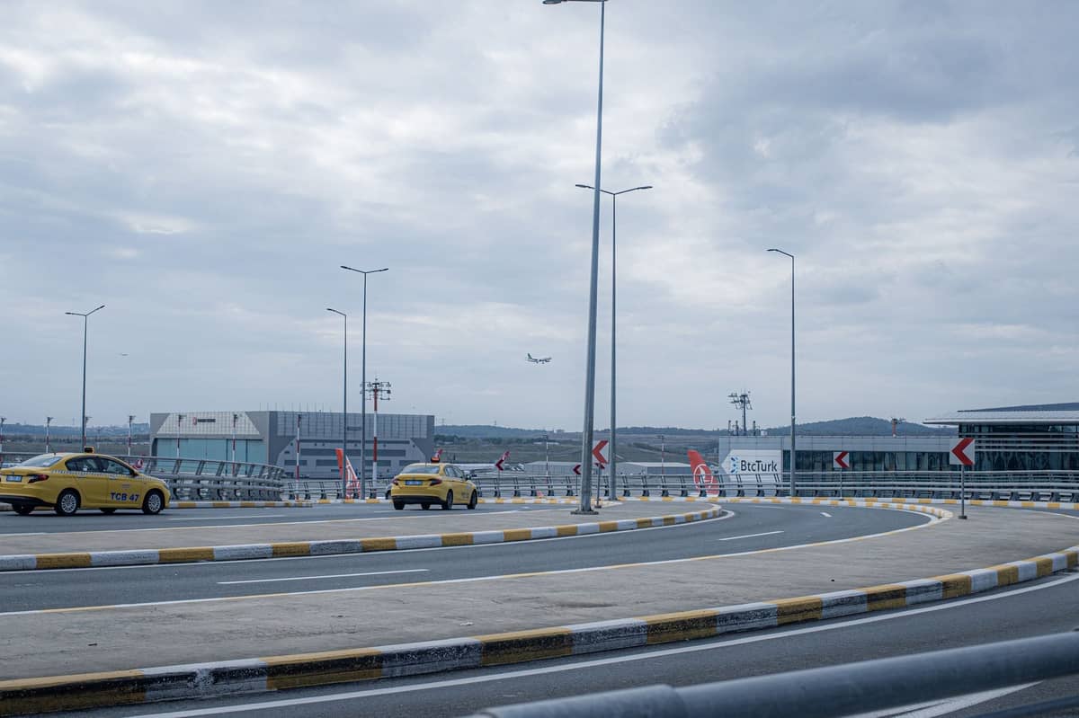 Bulutlu bir gökyüzü altında arka planda iki sarı taksi, havalimanı terminalleri ve kalkan bir uçakla havalimanına giden virajlı erişim yolu