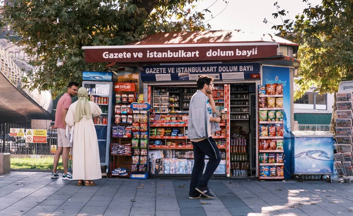 İstanbul'da bir sokak büfesi, Istanbulkart satışı ve bakiye yüklemesini tanıtan bir tabela ile insanlar alışveriş yapıyor ve yanından geçiyor.