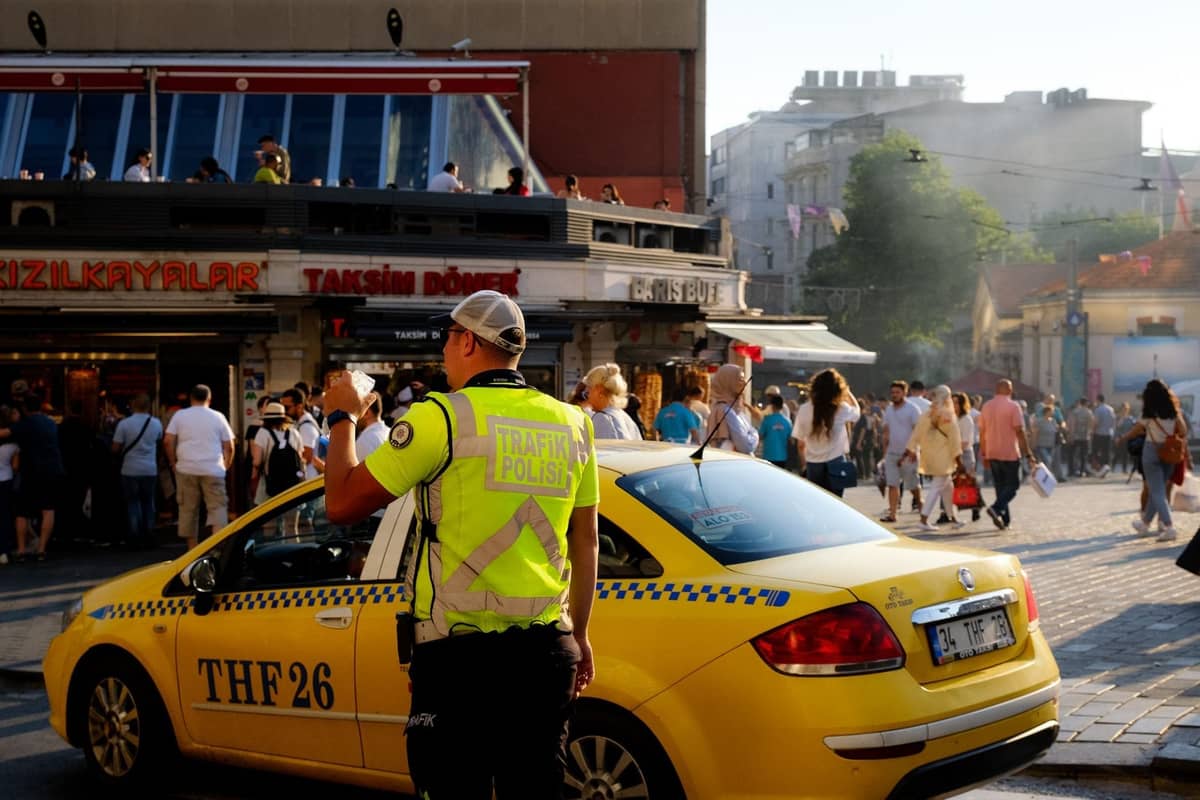 Kalabalık bir meydanda durmuş sarı bir İstanbul taksisi, yakınında trafik polisi ve Taksim Döner yazan dükkanların önünden geçen insanlar.