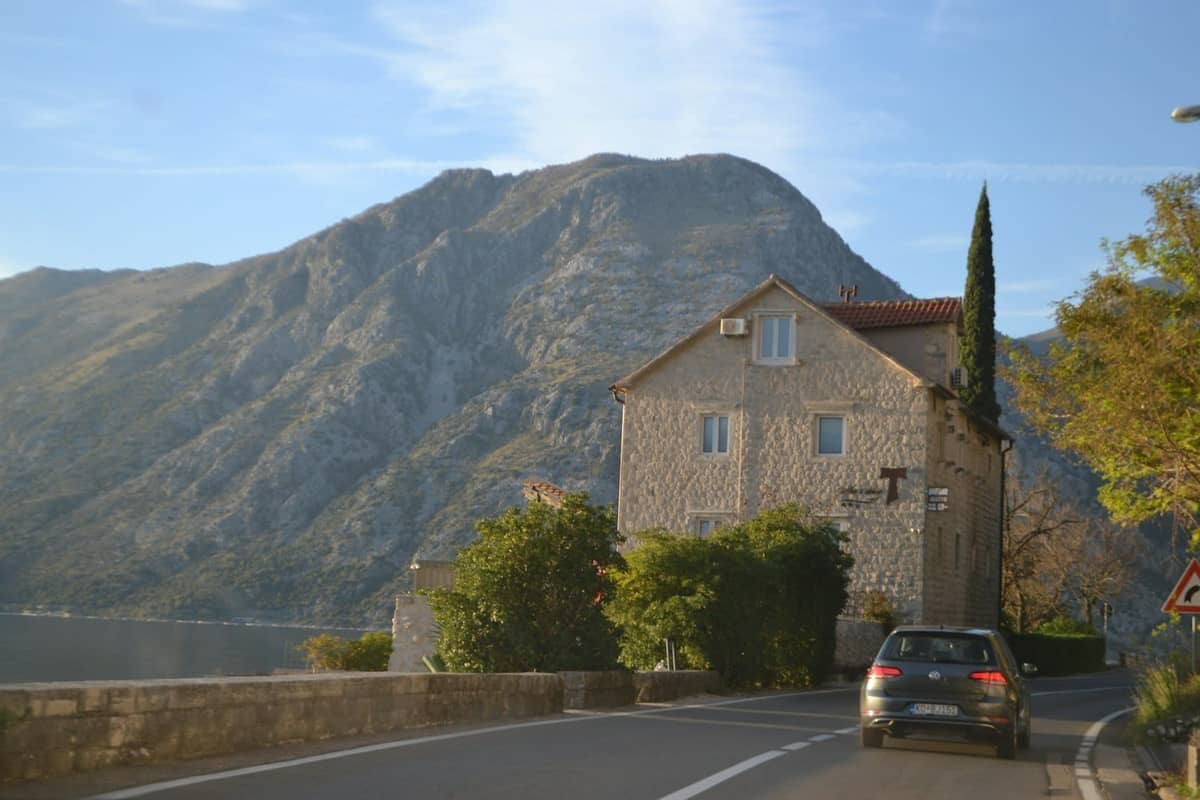 A car drives along a coastal road in Kotor, passing stone houses with a mountain backdrop and the bay visible nearby.