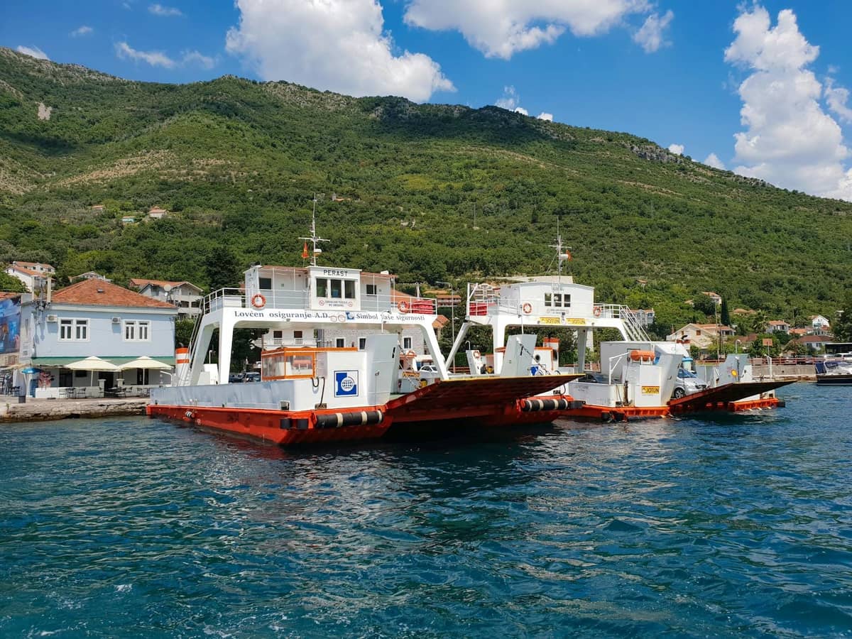 Car ferries docked at a harbor on the Bay of Kotor with green hills and buildings in the background.