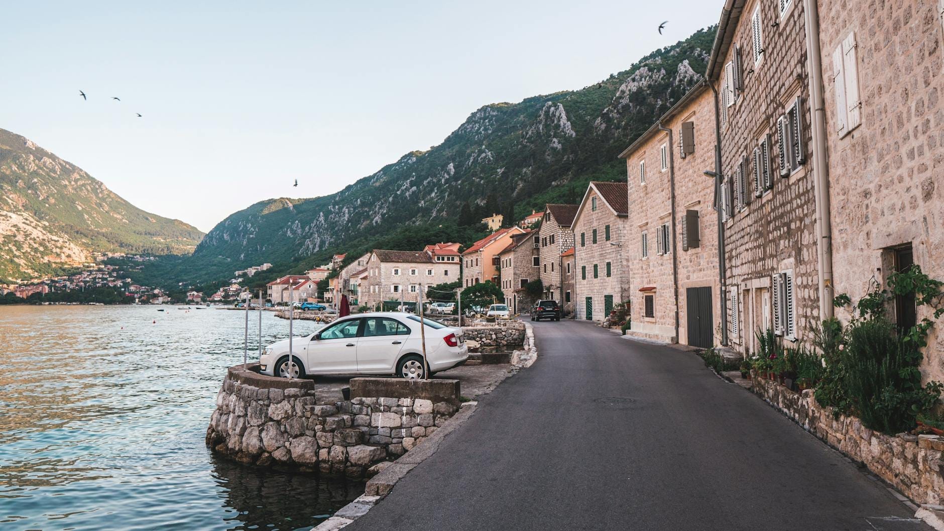 Edifícios de pedra alinham uma tranquila estrada à beira-mar na Baía de Kotor, com um carro estacionado perto da água e montanhas verdes ao fundo.